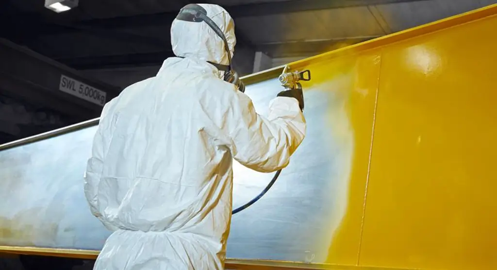 Arbeiter in Schutzkleidung beim Lackieren einer großen Metallplatte in Gelb.