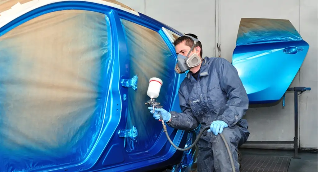 Autolackierer in Schutzkleidung sprüht blaue Farbe auf die Karosserie eines Fahrzeugs in einer Lackierkabine.