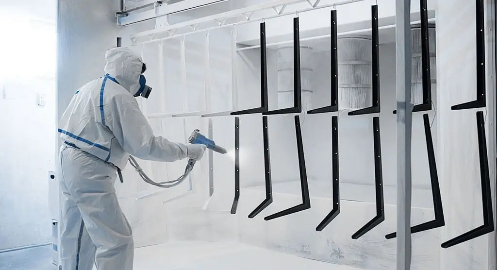 Arbeiter in Schutzanzug beim Pulverbeschichten von Metallrahmen in einer Kabine.