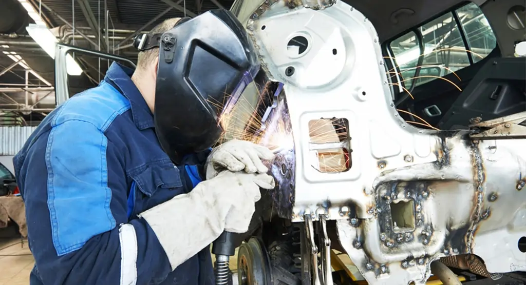 Arbeiter mit Schweißmaske, der an einem Fahrzeugrahmen Schweißarbeiten durchführt.