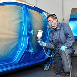 Autolackierer in Schutzkleidung sprüht blaue Farbe auf die Karosserie eines Fahrzeugs in einer Lackierkabine.