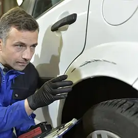 Mann im Blaumann inspiziert eine Autokarosserie auf Schäden.